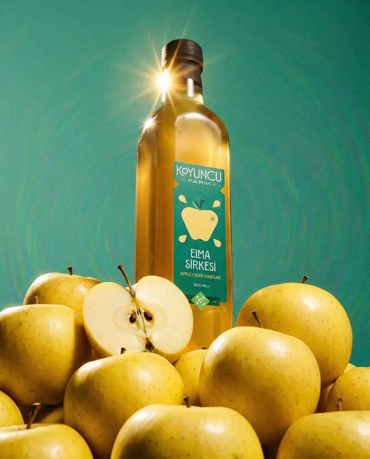 Vinegar bottle backlit against a teal backdrop above a pile of apples.